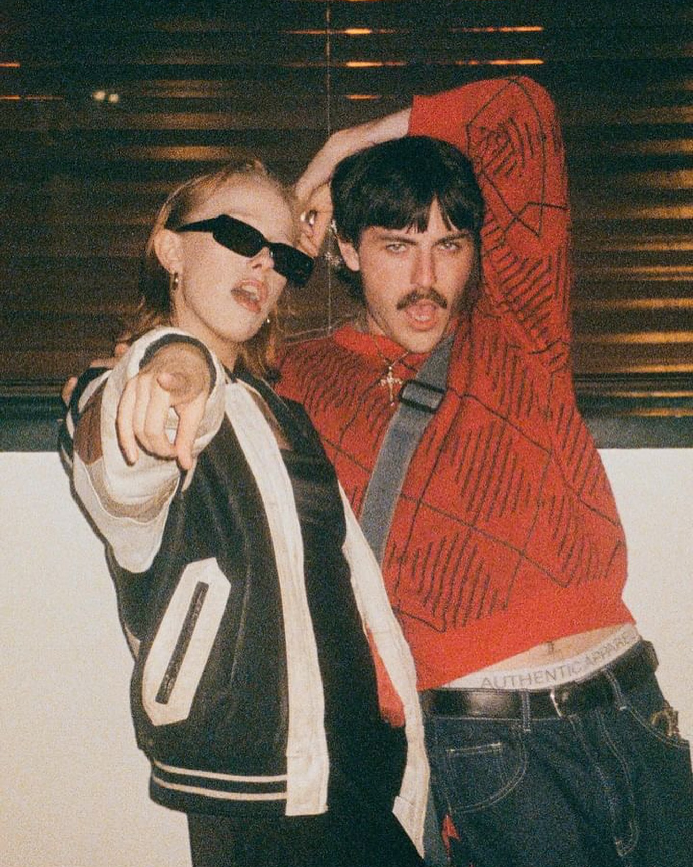 the image is a vintagestyle photograph, likely taken with film, showing a young couple.  the woman is wearing a black and white varsitystyle leather jacket, sunglasses, and is pointing directly at the camera. the man is wearing a red patterned sweater,  denim jeans, and has a mustache. they appear to be close, with the man's arm around the woman's shoulders. the background suggests a nighttime urban setting. the photo has a grainy, slightly outoffocus quality typical of older film photography, lending it a retro aesthetic. the overall style is very 90s influenced.