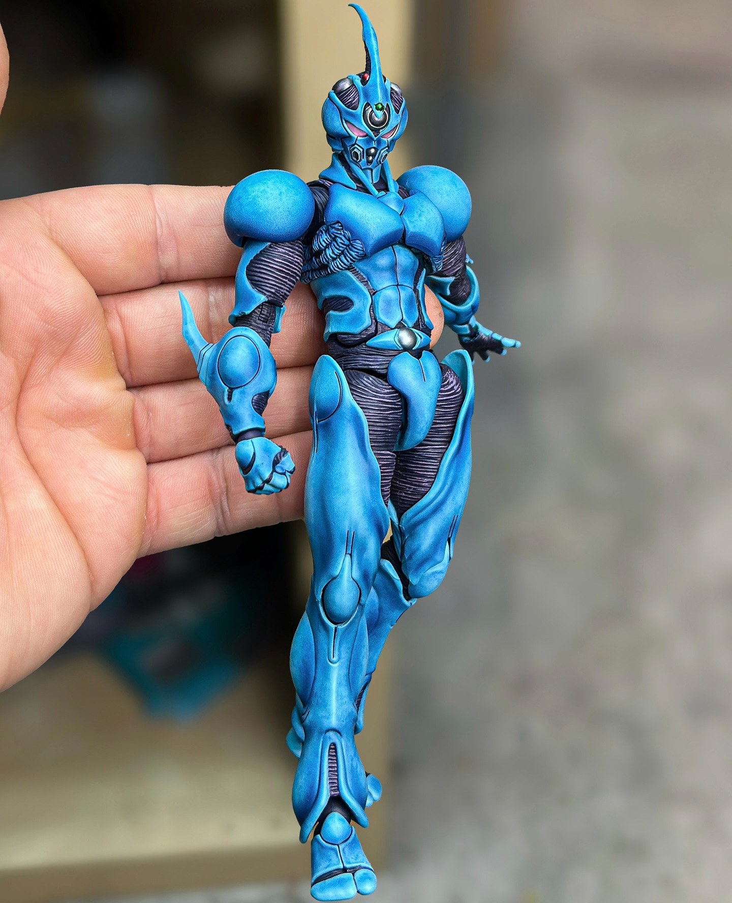 a hand holds a detailed, blue guyver action figure. the figure is painted with intricate detail and resembles an alien or bioboosted armor. the figure is likely a collectible toy.