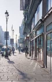 a cobblestone street in paris, france, with people walking on the sidewalk past shops and buildings. the sun is shining brightly in the morning, casting long shadows on the street. there are lampposts with streetlights along the sidewalk.