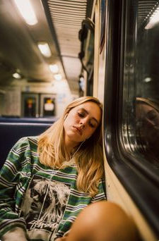 a young woman with long blonde hair is sleeping on a subway train. she is wearing a green and white striped sweatshirt and has earphones in. the lighting is dim and the atmosphere is calm. the photo is taken from eye level and captures the woman's reflection in the window.