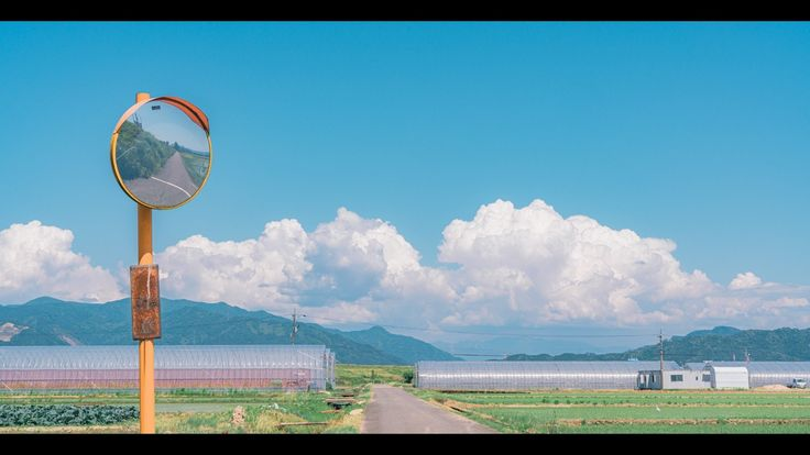 a scenic shot of a rural landscape with a road running through fields, flanked by greenhouses. in the foreground, a safety mirror reflects another part of the road, and a clear blue sky with fluffy clouds forms the backdrop, with distant mountains adding to the picturesque scene.