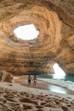 a couple is standing on a beach inside a large sea cave. the cave is carved out of the cliff face and opens up to the ocean. the couple is holding hands and looking out at the view.  the water is calm and clear, and the sand is white and powdery.  this looks like the algar de benagil cave in portugal.
