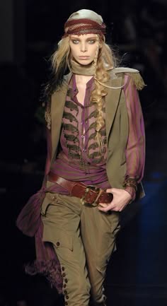 a woman wearing a stylish outfit poses for a fashion show on the runway. she is wearing a purple and green layered top, brown leather pants, a belt and a brown bandana. her hair is braided and she has natural makeup on.