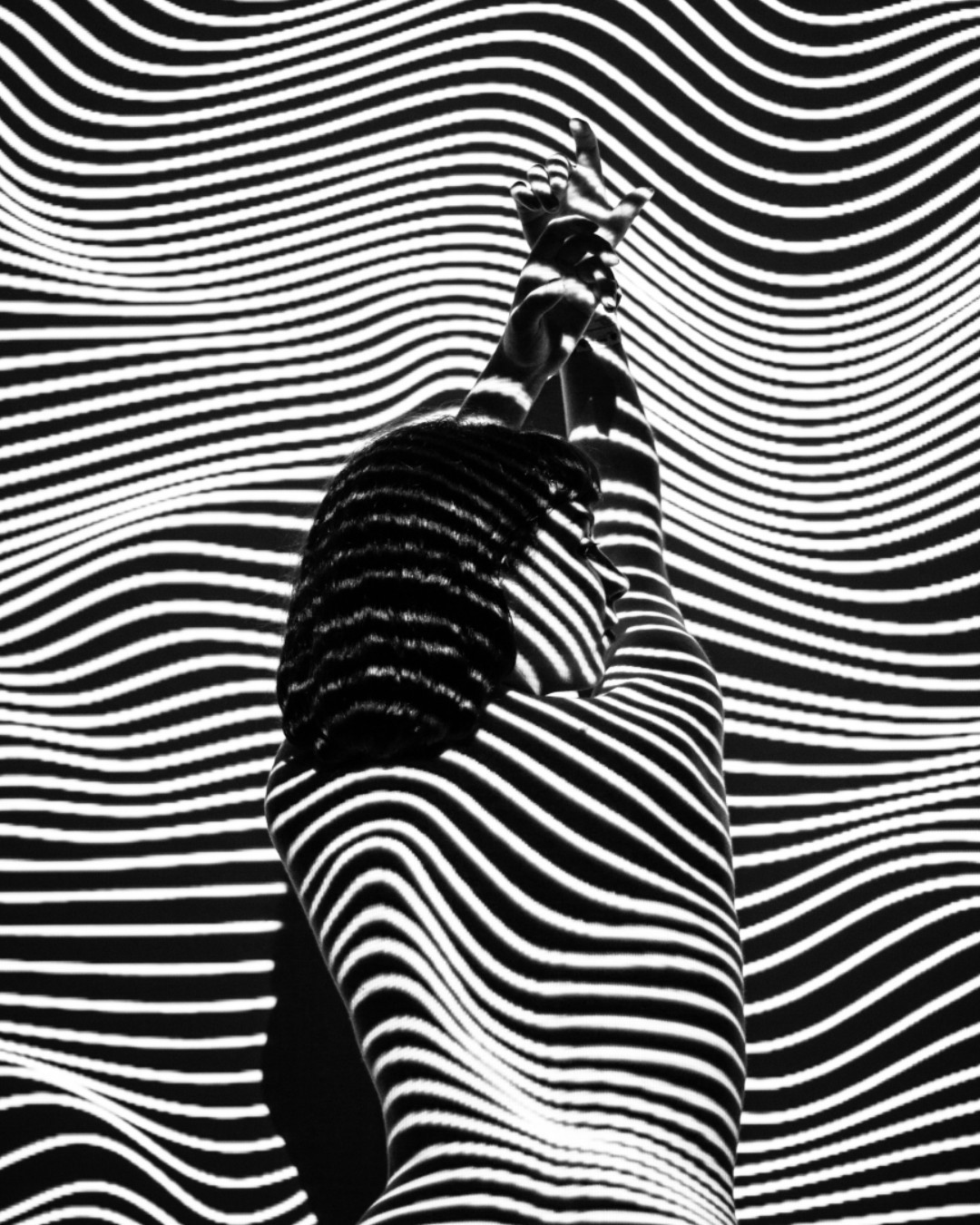 a black and white photograph of a woman's silhouette, posed with arms raised, bathed in a light pattern of intersecting stripes that forms a graphic and sensual abstraction. the image captures a sense of movement and dynamism, creating an intriguing interplay of light and shadow.