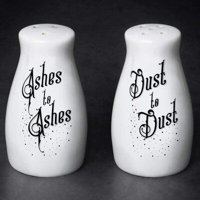 two white porcelain salt and pepper shakers sit side by side on a dark gray surface. the left shaker has "ashes to ashes" written on it in a decorative black font, with small black dots sprinkled around the words. the right shaker has "dust to dust" written in a similar style. the shakers have rounded shapes and a small set of holes in the top. they are novelty items with a humorous take on a common phrase.