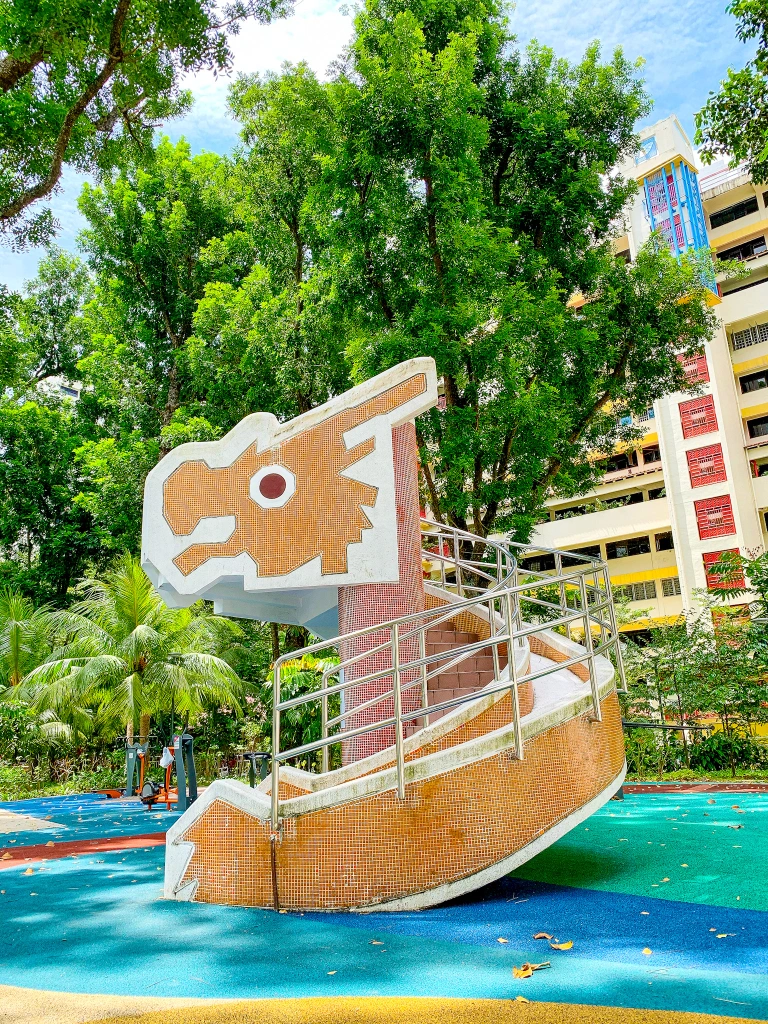 a brightly colored playground slide shaped like a dragon's head. the slide is made of concrete and has a metal railing. the slide is located in a park and there are trees in the background. the slide is surrounded by blue and green ground cover.