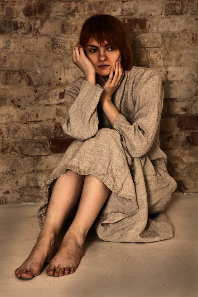 a woman with red hair and a light brown dress is sitting on the floor with a brick wall behind her. her hands are supporting her face, and she has a sad expression. she is barefoot. the overall impression is one of despair or poverty.