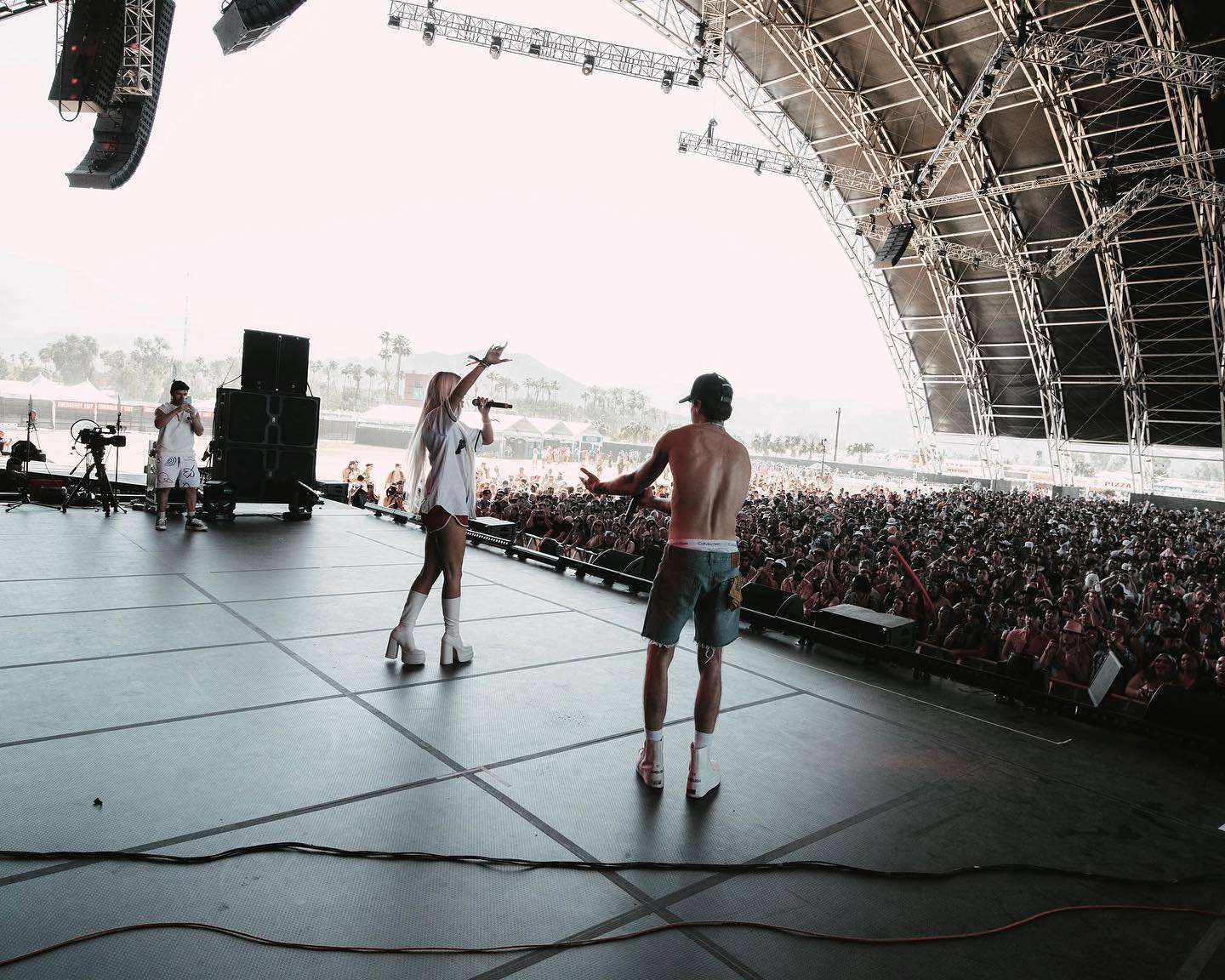 a wide shot of a music concert, showcasing a female singer holding a microphone on stage with a shirtless man next to her. they are performing in front of a massive crowd under a large stage canopy. there are speakers and other equipment on stage, along with a cameraman recording the performance. the audience stretches far into the distance, showing the scale of the event.