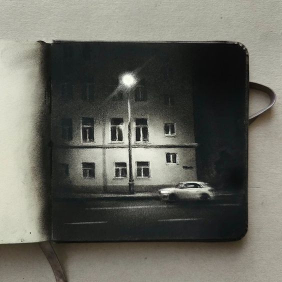 the image showcases a black and white drawing or illustration depicting a quiet, nighttime city scene. the focus is on a multistory apartment building under a single streetlight. a car is parked or slowly driving along the street in front. the style is realistic, with attention to detail in the building's architecture and the soft shadows created by the nighttime lighting. the overall mood is serene and slightly melancholic. the art is presented within an open sketchbook, indicating a work in progress or a finished piece displayed in its original medium. the dark tones and grayscale palette contribute to the overall atmospheric feel.
