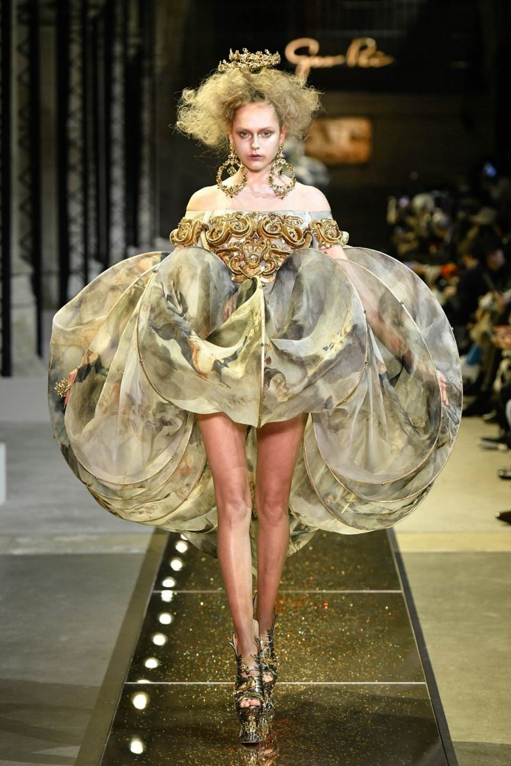 a model walks the runway wearing a breathtaking haute couture gown. the dress is offtheshoulder, with a fitted bodice heavily embellished with gold baroqueinspired detailing. the skirt is voluminous and made of layers of sheer fabric printed with a renaissancestyle painting. the model's hair is styled in a large, curly updo, and she wears ornate gold earrings and a gold crown.  she is wearing high platform heels that complement the overall opulent aesthetic. the runway is gleaming and has subtle lighting accents. the background is blurred, suggesting a high fashion show setting. the overall impression is one of grandeur and artistic expression.