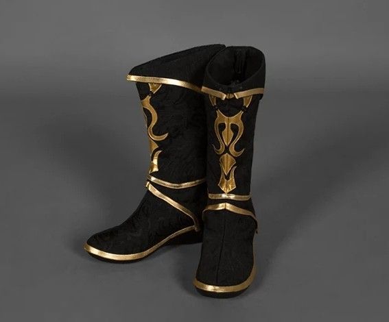 the image shows a pair of black kneehigh boots with gold decorative trim. the boots appear to be made of a soft fabric material with gold accents along the soles, straps, and ornate designs on the upper part of the boots. one boot is slightly angled back while the other is facing forward. the background is a muted grey color.