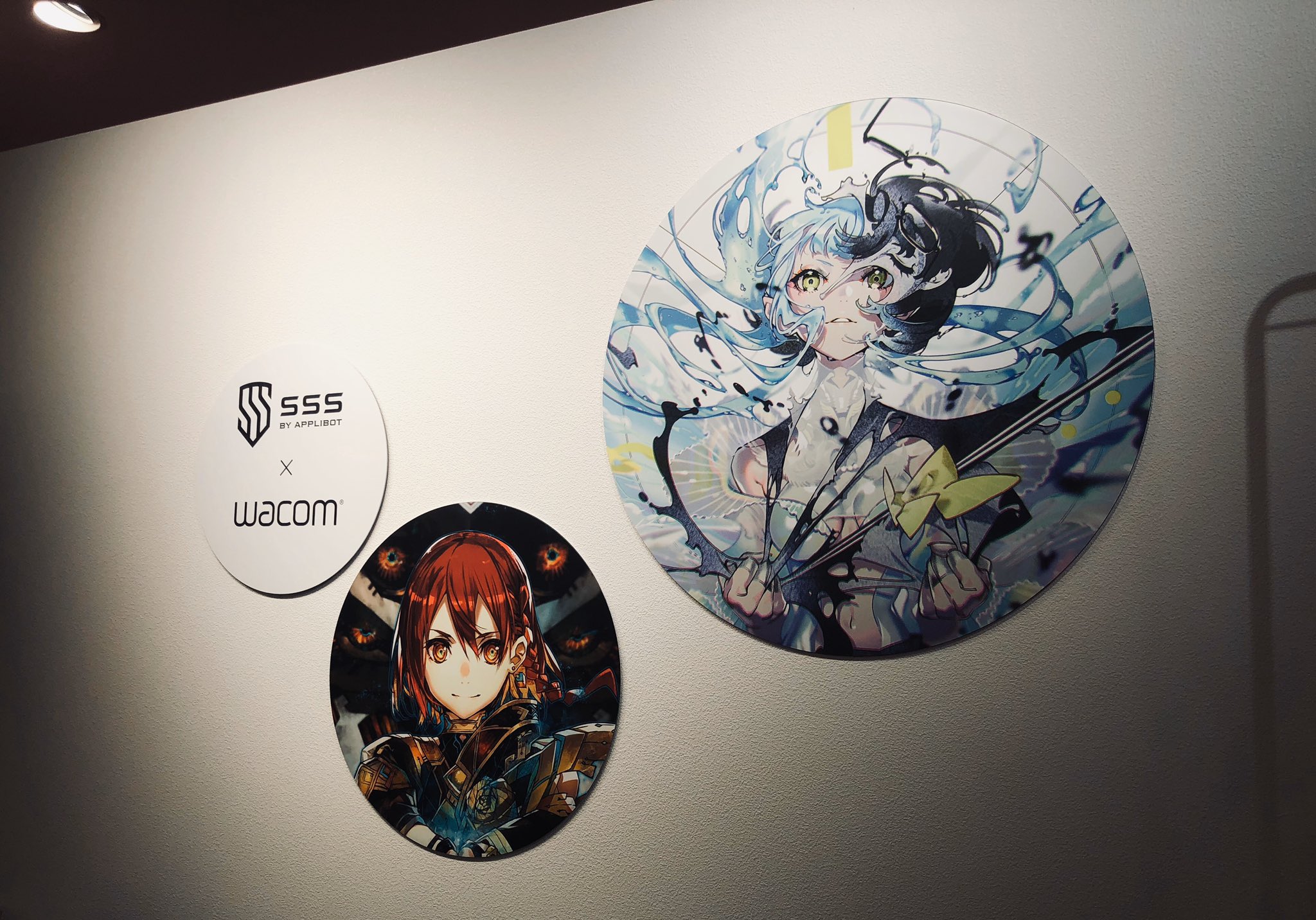 the image shows an indoor wall featuring promotional art for a collaboration between the mobile game "sss by applibot" and wacom. two large circular posters showcase animestyle characters, likely from the game. the upper poster features a girl with blue and black hair wielding a sword, while the lower one depicts a girl with red hair in armor. a smaller sign between them announces the collaboration, displaying the logos of both "sss by applibot" and "wacom". the wall is white, and the lighting appears to be artificial, likely from ceiling fixtures.