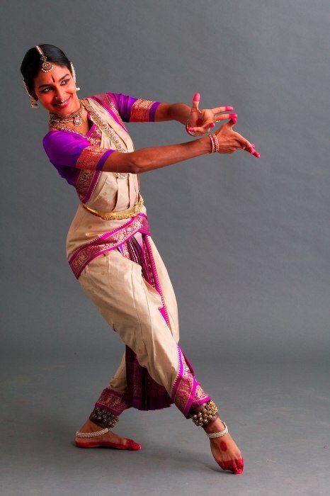 a young indian woman in a traditional sari, adorned with ornate jewelry, performs a graceful dance pose in a studio setting. her expression is focused and her hands are in a delicate gesture, while her feet are poised in a captivating stance. the woman's attire and movements are both elegant and vibrant, reflecting the rich culture of indian classical dance. the backdrop is a simple grey, allowing the dancer to take center stage and showcase her artistry.