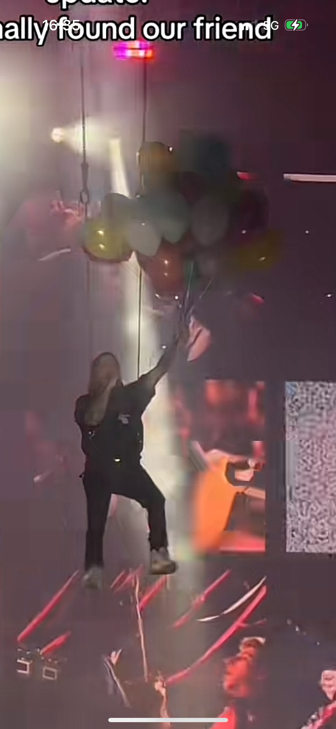 a blurry shot of a rock concert with people in the background, and a singer in the foreground with balloons hanging above. the singer is holding a microphone and appears to be performing on stage in front of a crowd of excited concertgoers. the image is dark with spotlights illuminating the stage.
