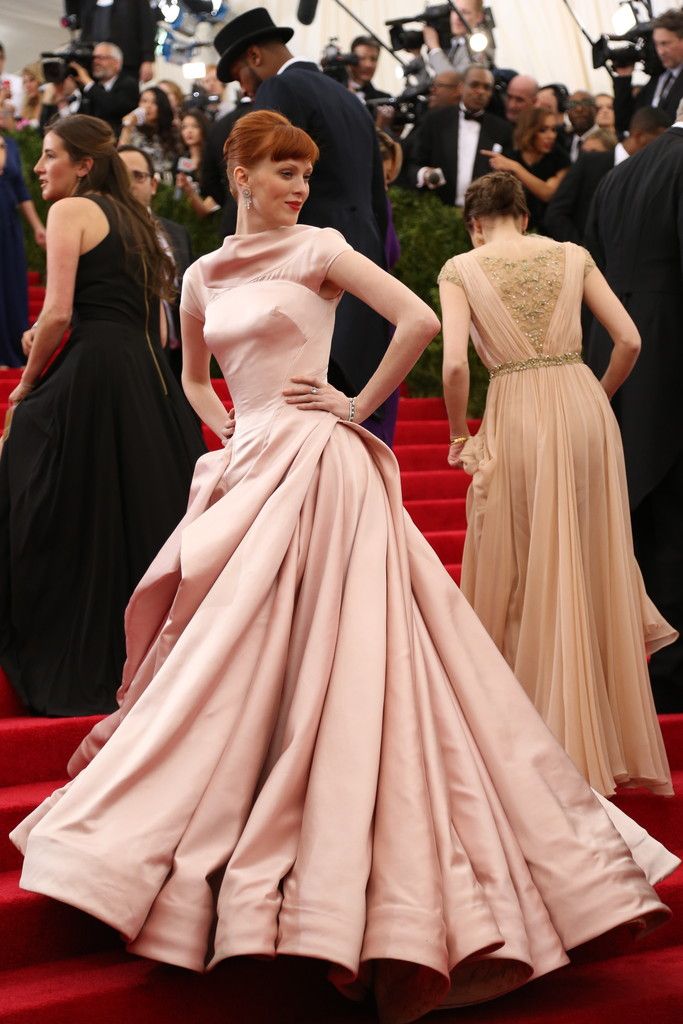 a woman in a long pink gown stands on a red carpet with a crowd of people behind her. the woman is smiling and appears to be posing for the camera. there are other women in dresses visible in the background.