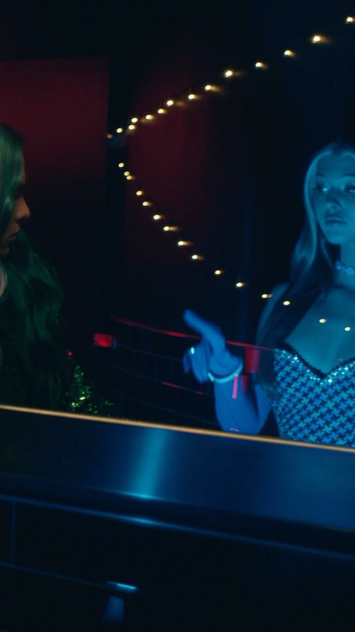 two women are at a bar in a nightclub with neon lighting and string lights in the background. one woman has green hair, and the other is wearing a houndstooth patterned dress.