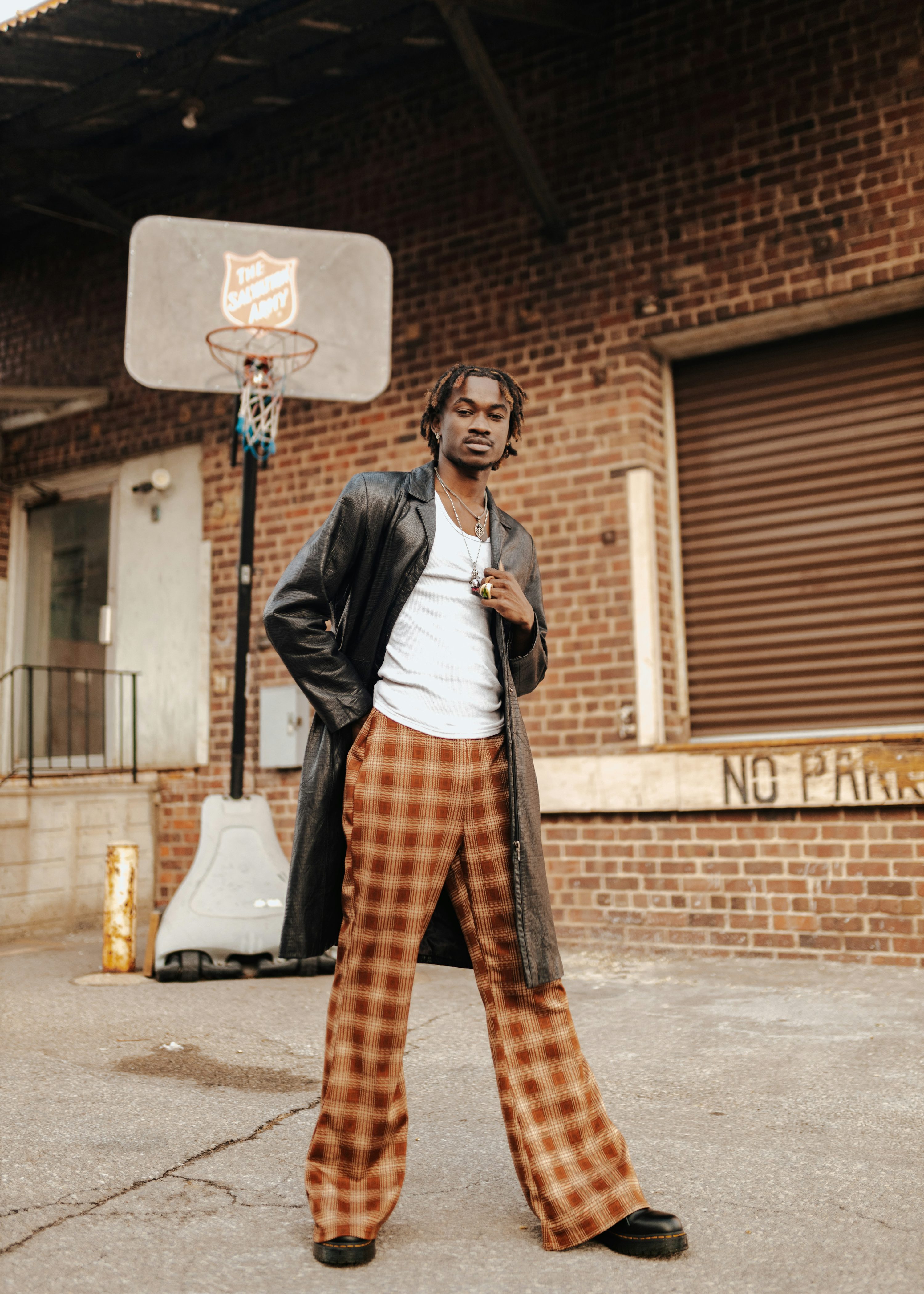 a stylish young black man stands confidently in front of a brick building with a basketball hoop. he's wearing a long black leather coat, a white tshirt, and brown plaid bellbottom pants. he's accessorized with a necklace and ring, and his footwear is black doc martens. the overall setting and the model's attire suggest a blend of urban and vintage fashion styles.