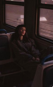 a young woman with dark hair and a fringe is sleeping on a train. she is wearing a darkcoloured jacket and trousers, and her head is resting against the back of the seat. the lighting in the carriage is low, with the warm glow of sunset coming through the window. the image evokes a sense of peace and solitude, capturing the quiet moments of a long journey.