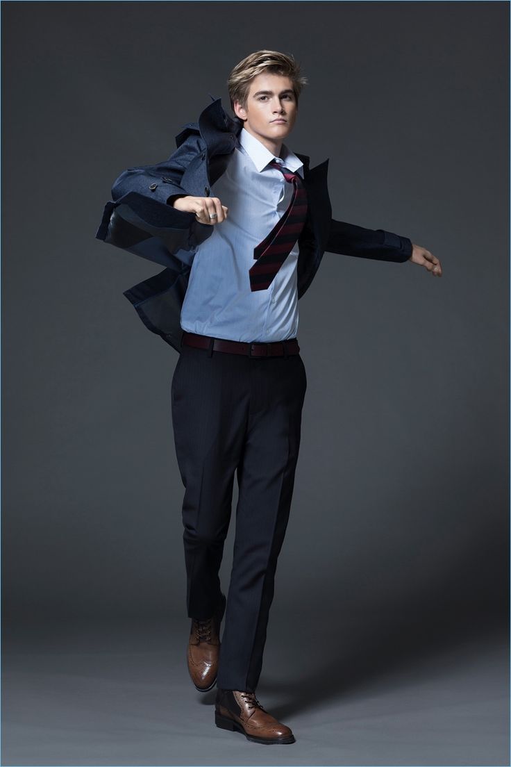 a young man in a blue suit and a white shirt stands against a gray background, holding his jacket open and posing with one foot in the air. he is wearing a striped blue and red tie, black pants, and brown shoes.
