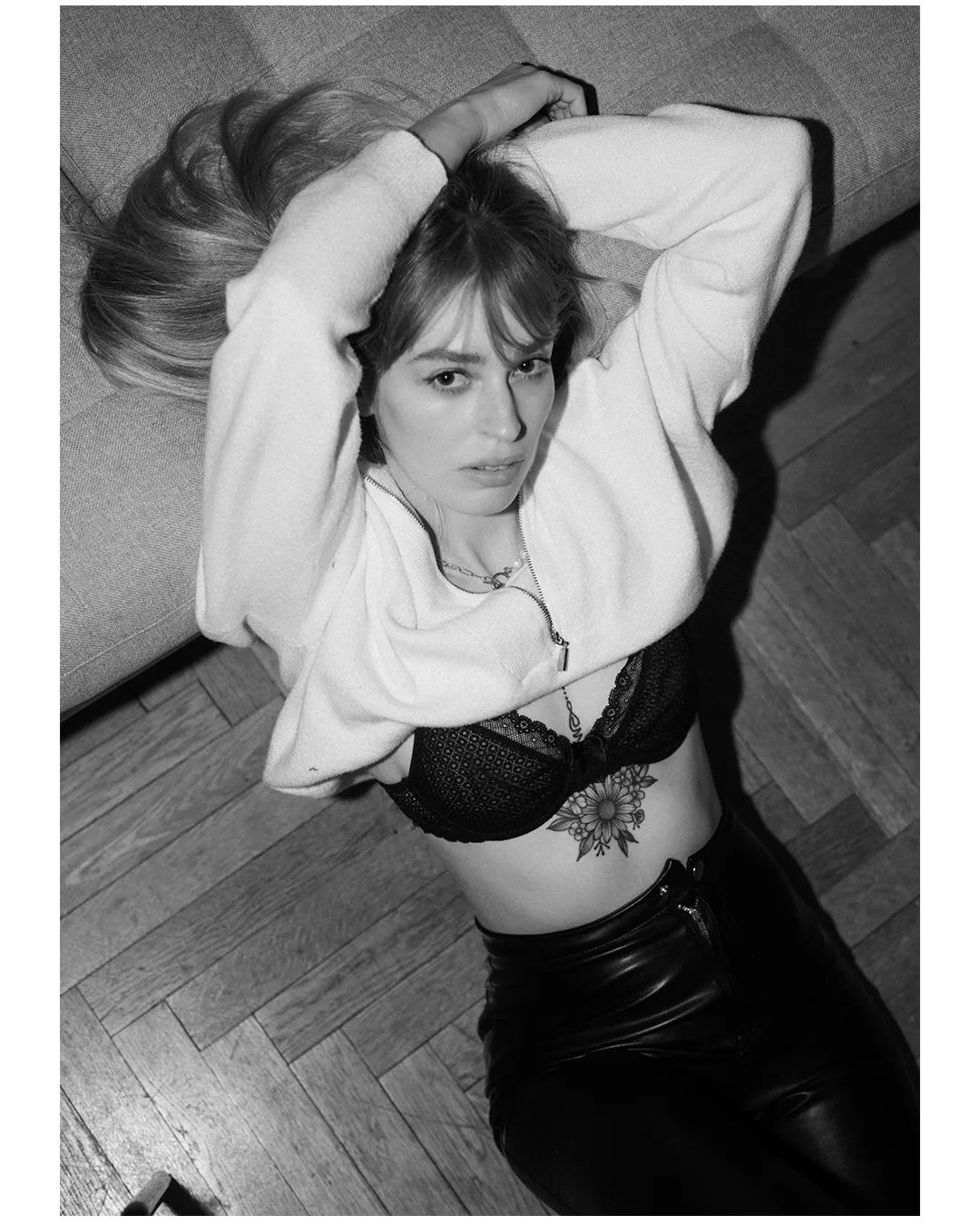 a young woman in a white sweater, black bra, and leather pants sits on a couch, looking at the camera. her hair is long and blonde, and she has a tattoo on her stomach. the photo is in black and white.