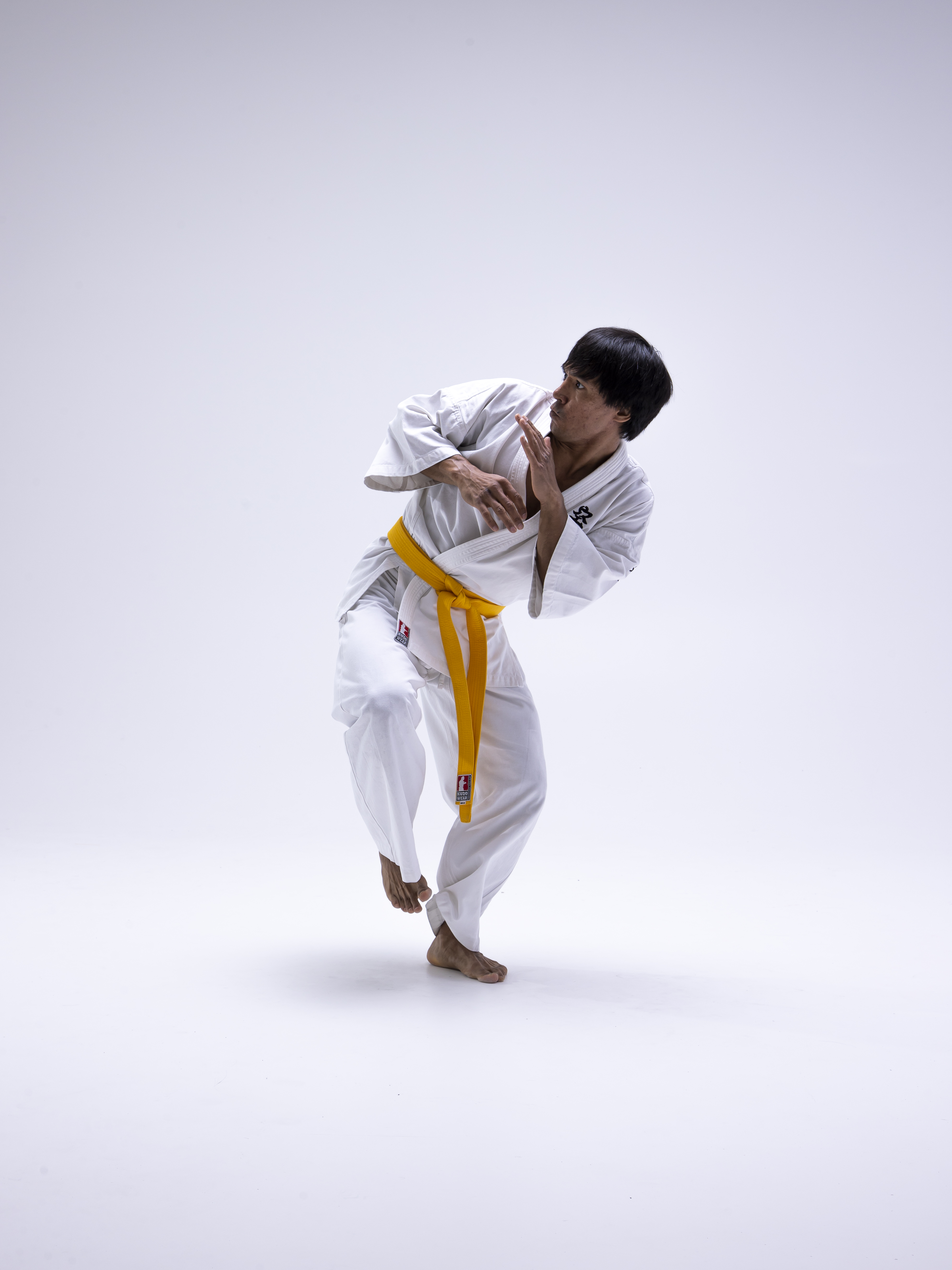 a man dressed in a white karate uniform with a yellow belt is shown in a defensive stance against a white background. he is barefoot and his hands are up in a blocking position, looking to his left.