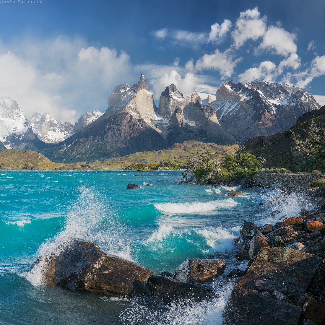 the image captures a breathtaking view of a vibrant blue lake with waves crashing against a rocky shore. in the background, majestic snowcapped mountains rise up into a sky dotted with white clouds. the sun illuminates the scene, casting shadows on the rocks and highlighting the turquoise hues of the water.  the overall impression is one of aweinspiring beauty and the raw power of nature.