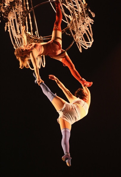 two female aerial artists are performing a gravitydefying act. one is hanging upside down from an aerial silk attached to a chandelier, while the other hangs by her feet from another silk. they are both gracefully posing with their bodies contorted in a display of strength and flexibility. the black background highlights the beauty and skill of the performers. the scene evokes a sense of wonder and excitement, typical of a circus or theatrical performance.