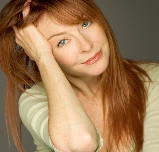 a woman with long red hair is looking down and to the side. she is wearing a lightcolored shirt and has a casual, relaxed look. she is in a studio setting with a grey background and is wearing light makeup. her skin is clear and bright, her hair is long and flowing and her eyes are a clear green.