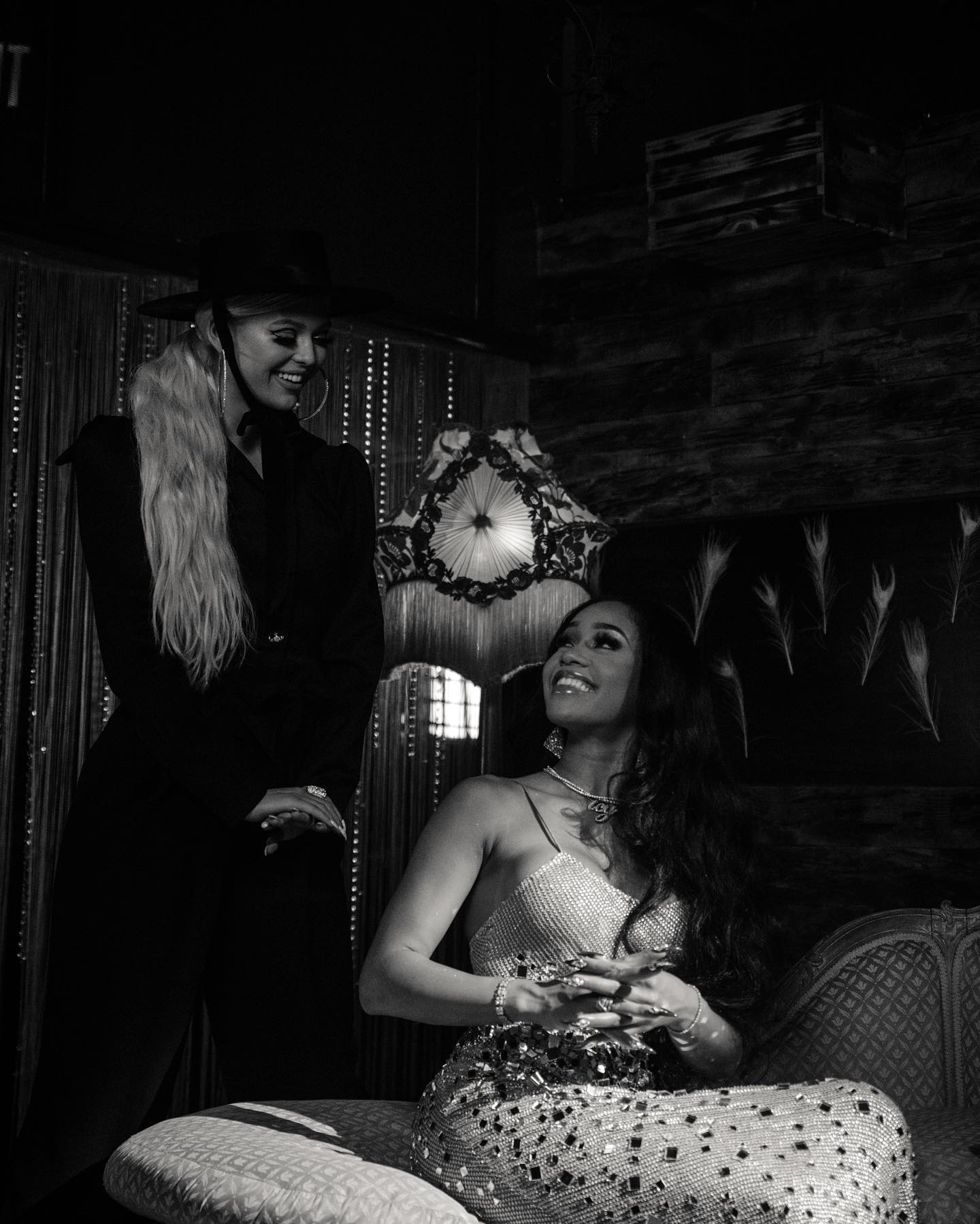 two women are pictured in a black and white photograph. one woman is standing and wearing a hat and a dark suit, and the other is sitting on a bed in a sparkly dress. they are both smiling and looking at each other. there is a lamp behind them and feathers on the wall.