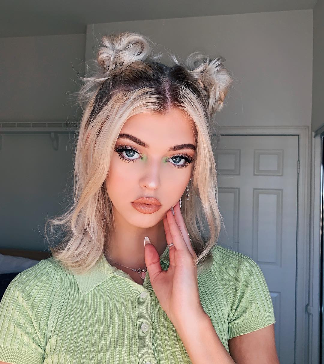 a portrait of loren gray wearing a light green collared shirt with blonde hair styled in space buns. she has green eyeshadow on and is wearing a necklace and a ring. her hand is resting on her face. a white door is visible in the background.