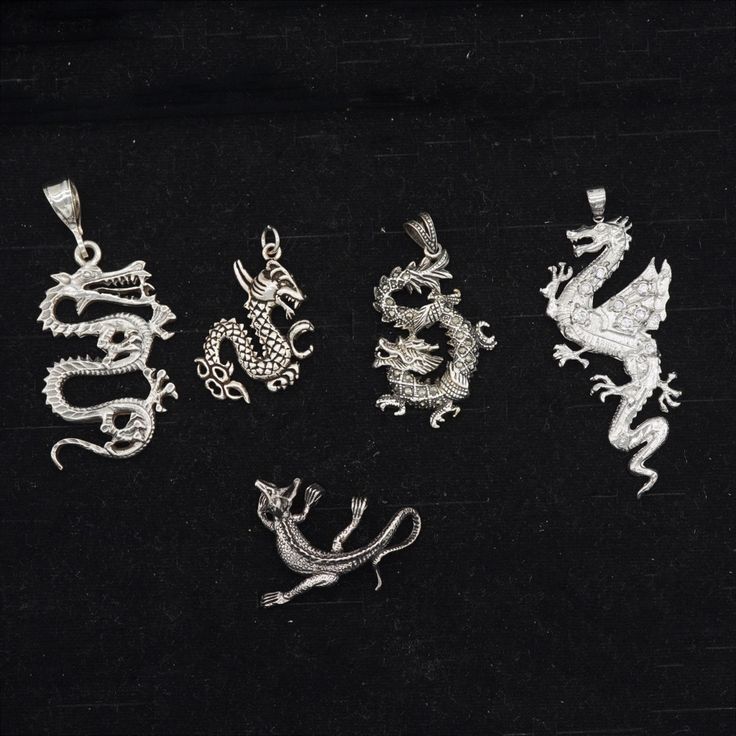 the image contains a collection of five silver dragon pendants. they are arranged on a black surface. the designs vary from intricate, coiled dragons to winged dragons.