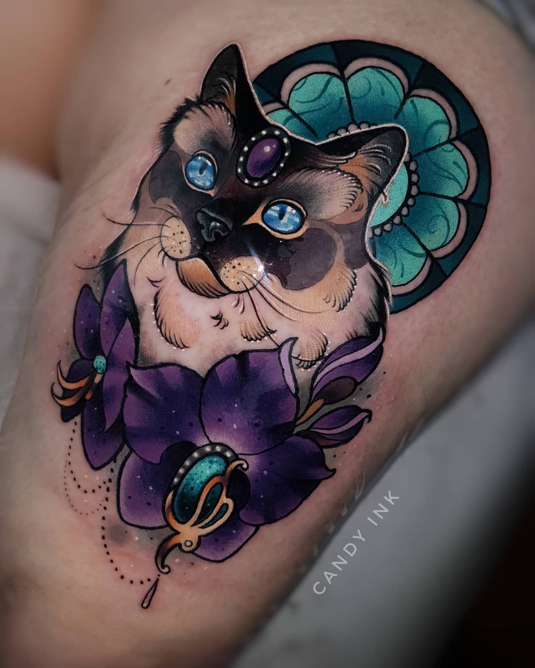 a person's upper thigh with a neotraditional style tattoo of a siamese cat. the cat is the focal point with bright blue eyes. it has a purple jeweled ornament on its forehead. behind the cat's head is a turquoise and teal stained glassstyle design. below the cat's face are two, large, purple orchids. the orchids also have a green jeweled ornament with gold detail. the tattoo is detailed and colorful with black line work and white highlights. the artist's signature "candy ink" is written in cursive script below the orchids.