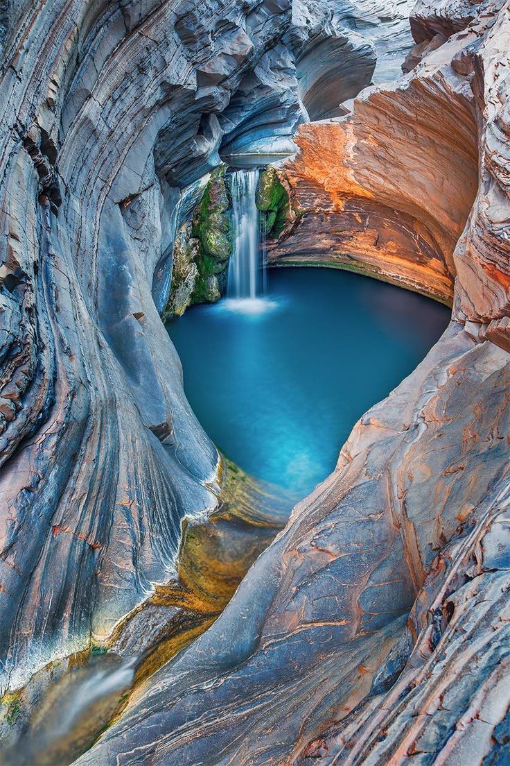 a waterfall cascades into a pool of crystal clear blue water nestled in a canyon of layered rock. the surrounding rock faces are a mixture of gray, brown, and orange hues, creating a striking contrast with the vibrant blue water. the scene is a testament to the beauty of nature's sculpting power, showcasing a secluded paradise amidst the rugged landscape.