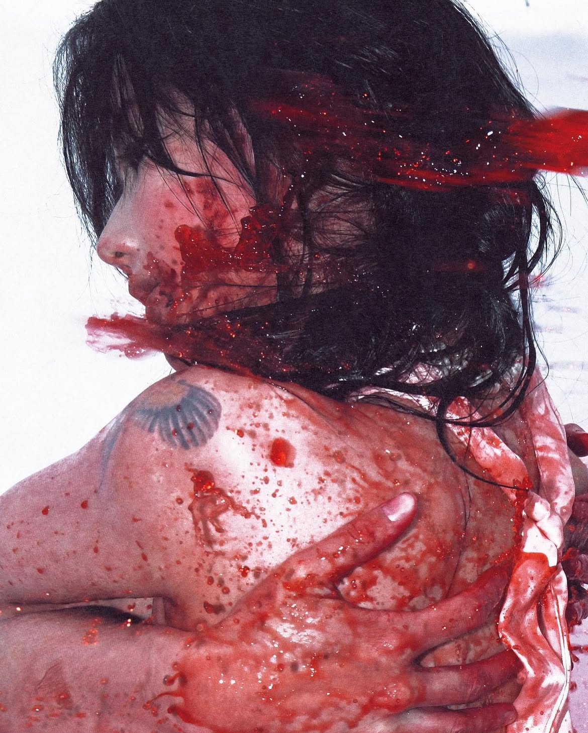 the image shows a woman with black hair covered in blood. blood splatters cover her face, neck, shoulders, and back. her hands, also covered in blood, are clasped around her back. a tattoo is visible on her left shoulder. the image is graphic and unsettling, depicting violence and gore.