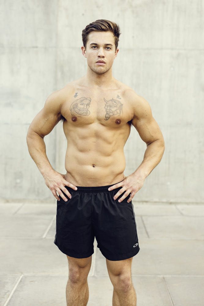 a shirtless man with a muscular physique and tattoos on his chest stands in front of a grey concrete wall. he is wearing black athletic shorts. he is looking at the camera. he is standing with his hands on his hips. he is posing for the camera.