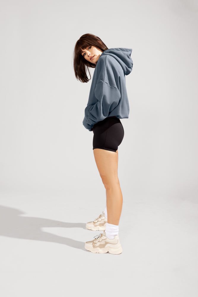 a woman is posing against a light grey background in an oversized blue hoodie, black biker shorts, white socks, and beige sneakers. she is facing sideways with her head turned towards the camera, and her hands tucked into the front pocket of her hoodie. the lighting is bright and artificial, creating soft shadows around the woman.