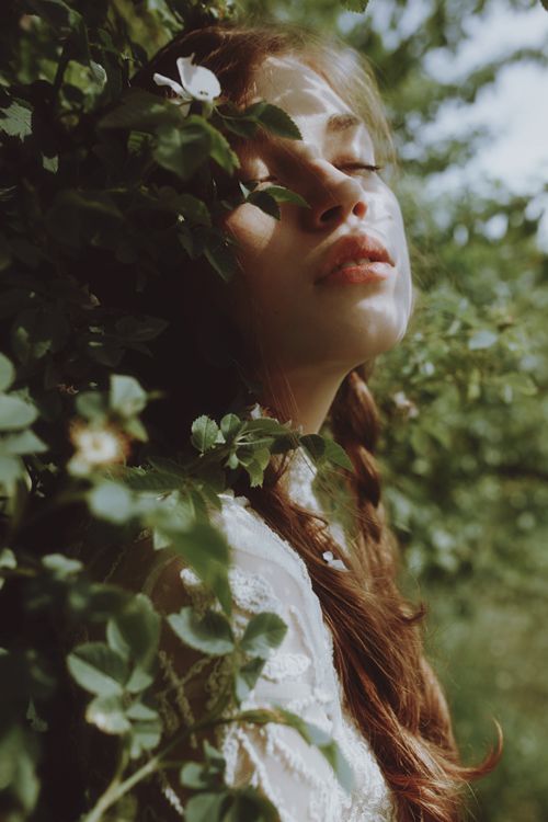 the image shows a young woman with long brown hair partially braided, her eyes closed, leaning against a backdrop of lush green foliage. she wears a white lace top that peeks out from the leaves.  sunlight filters through the leaves, casting a dappled light on her face and creating a serene and ethereal atmosphere.
