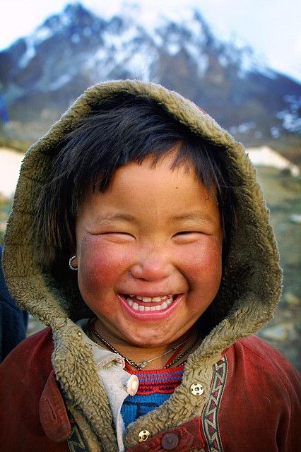 a closeup portrait shows a smiling child in a furlined hood, likely a young girl with bright, rosy cheeks and squinted eyes from laughter. she wears a red jacket with a patterned collar and has a necklace visible. the backdrop features a snowy mountain range, suggesting a winter environment.