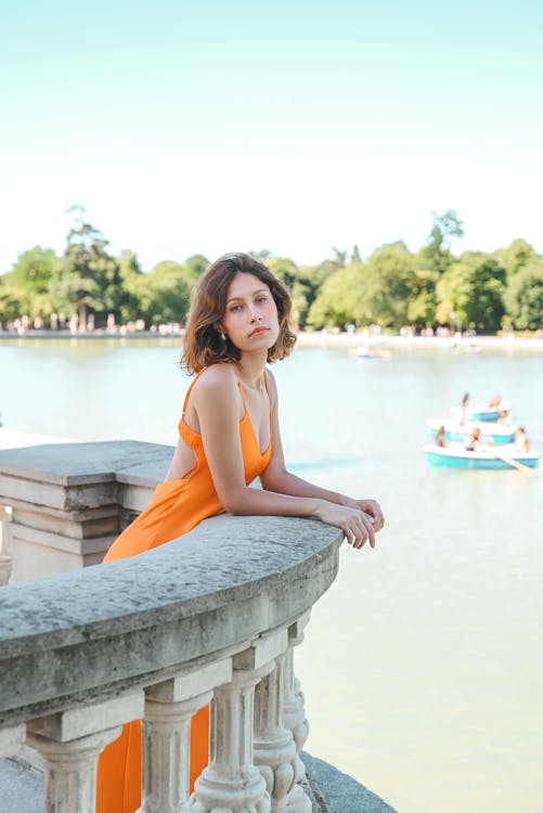 a woman in an orange dress is standing on a balcony overlooking a lake with trees in the background. she is leaning on the railing and looking at the camera. there is a boat on the lake with people in it. the photo was taken in el retiro park in madrid, spain. it is a sunny summer day.