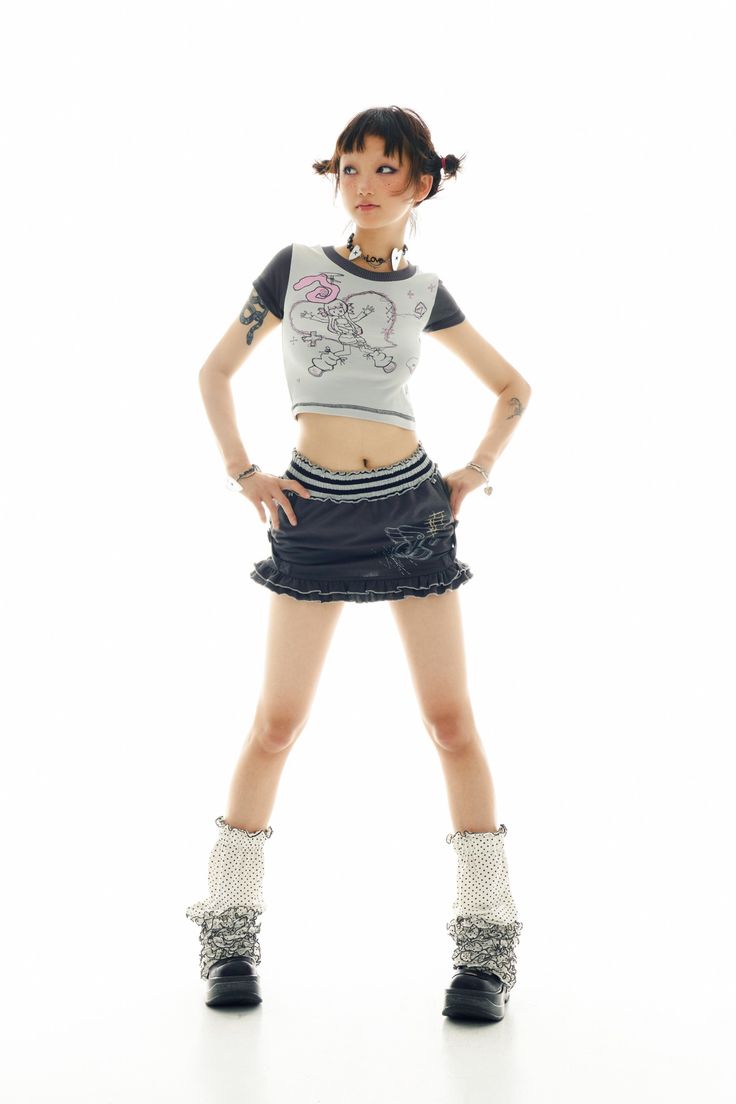 a young asian woman stands against a white background in a stylish outfit.  she's wearing a grey crop top with a cartoon design, a dark denim mini skirt with ruffles, patterned socks and chunky black boots.  her hair is styled with bangs and two small pigtails. she's looking over her shoulder with a slightly playful expression. the overall style is alternative, edgy, and trendy.