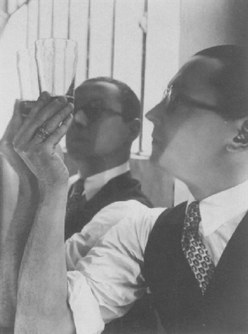 a black and white closeup photograph depicts two men, possibly from the 1920s or 1930s. one man is holding a glass up to the light, while the other man is looking over his shoulder. both men are dressed in formal suits and ties. the image is sharply focused and shows attention to detail, capturing the texture of the clothing and the expressions on the men's faces.  the setting appears to be indoors, perhaps in a home or office, and there are window bars visible in the background. the overall atmosphere is one of elegance and sophistication.