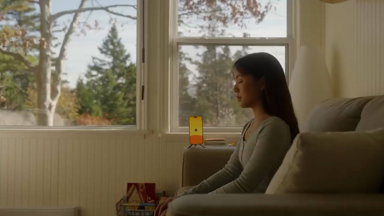 a woman is sitting on a couch in a living room. she is looking down at a smartphone that is propped up on a stand. the phone is displaying a yellow screen. there is a window behind her with a view of trees. there is a basket of books on the coffee table in front of her. the lighting is natural and the perspective is eye level.