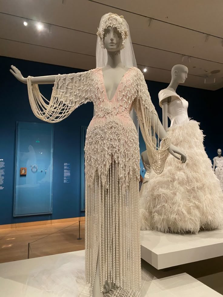 the image shows a wedding dress on a mannequin in a museum exhibit. the dress is covered in pearls and has a deep neckline. the mannequin is wearing a headpiece and veil. in the background, there is another dress on a mannequin and artwork on the wall.