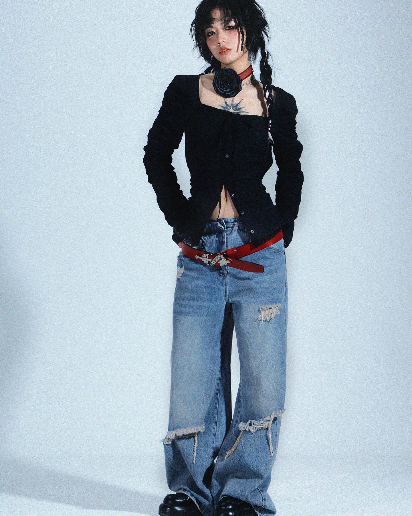 a young asian woman is posing in a studio against a light gray background. she is wearing a black squareneck longsleeved blouse with puffed sleeves, distressed light blue wideleg jeans, a red belt, and black boots. she has a black flower choker around her neck and her hair is styled in braids. she also has a tattoo on her chest. the overall style is dark, edgy, and fashionable.