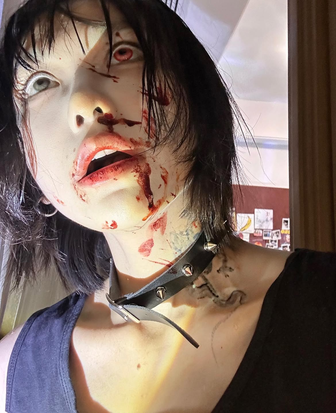 a closeup shot of a person with dark hair and pale skin, heavily made up to appear gory. the person has blood smeared around their mouth, nose, and on their neck. they wear a black choker with spikes. one eye appears to have a red contact lens, while the other is white. the person is wearing a black shirt. there is a tattoo on their neck.
