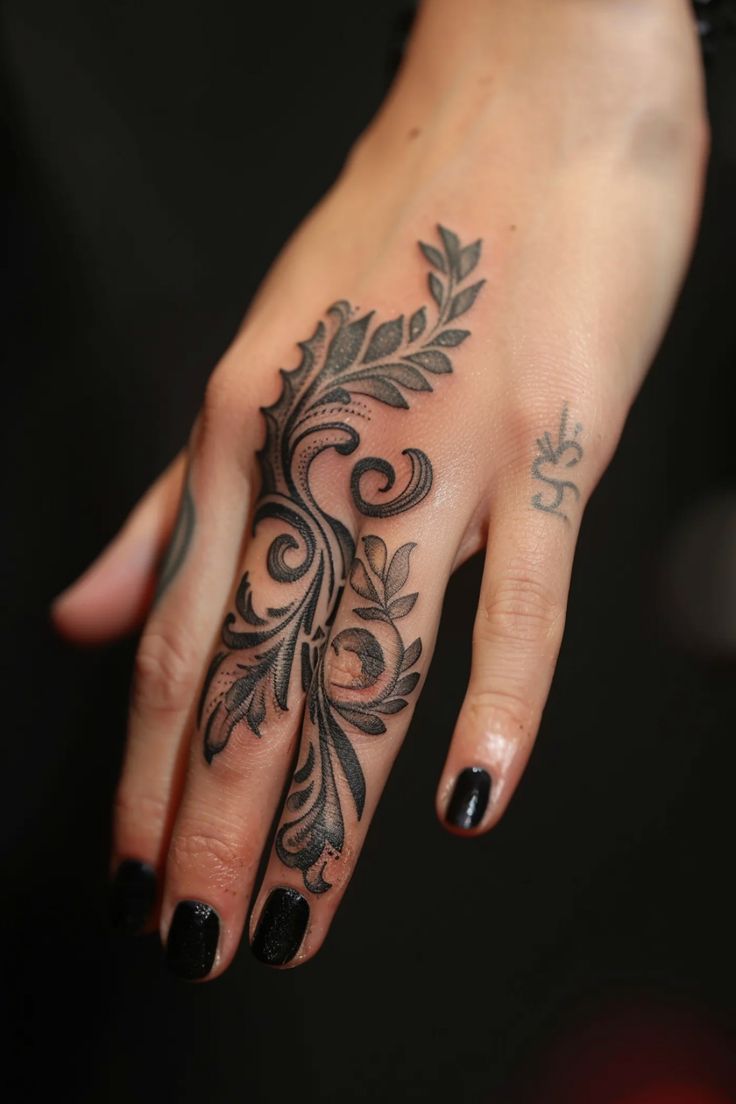 a closeup shot of a person's hand with an intricate black tattoo. the tattoo is located on the top of the hand and extends onto the middle finger. the design is ornate, featuring swirling patterns and leaflike shapes. the person is wearing black nail polish.
