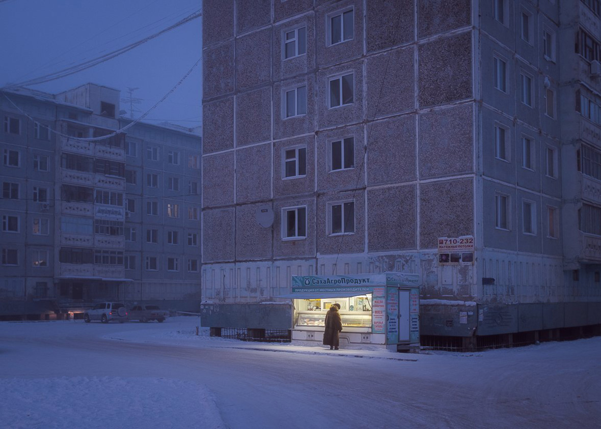 a snowcovered street in a russian city on a cold winter night. a small shop, brightly lit, stands on the corner. a lone figure walks past, shrouded in the darkness. the buildings on either side of the street are tall and imposing, with rows of windows reflecting the faint light of the street lamp. the atmosphere is quiet and desolate, with the snow falling softly and a chill in the air.