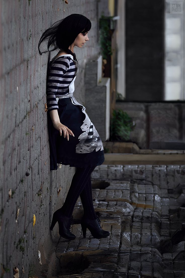 the image is a moody, atmospheric portrait of a young woman cosplaying a character, possibly from an anime or video game. she's standing in a narrow alleyway, leaning against a weathered brick wall. she has short, dark hair and is wearing a striped shirt and a dark, patterned skirt. her style is dark and somewhat gothic.  her pose and expression are pensive, giving a sense of quiet contemplation or solitude. the dark lighting and the grungy urban setting contribute to the overall mysterious and atmospheric feel of the photograph. the focus is clearly on the cosplayer, with the background slightly blurred.  the overall aesthetic is stylish and edgy.
