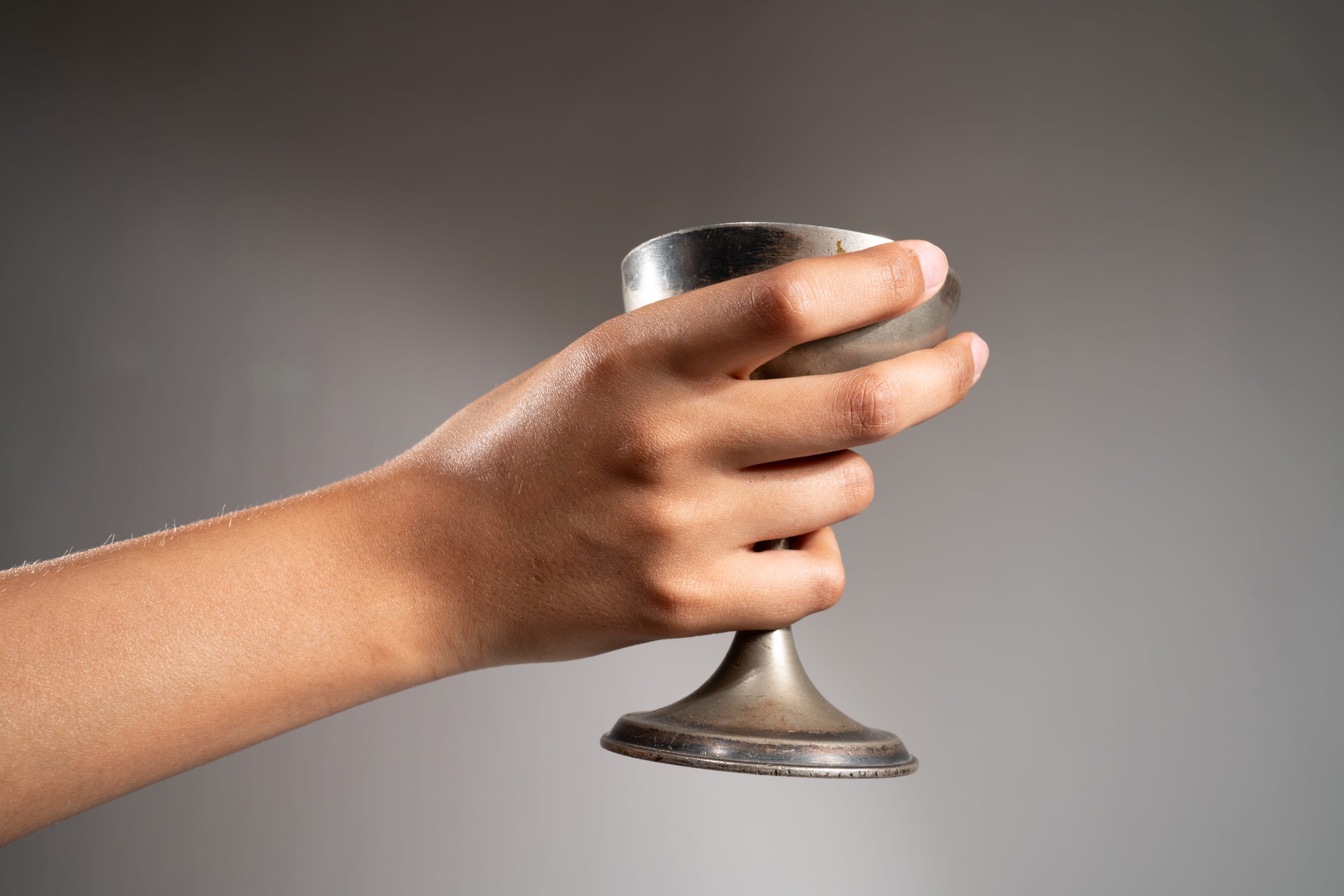 a hand is holding a silver metal cup against a light gray background. the cup is slightly tarnished and has a pedestal base. the hand is holding the cup securely, with the fingers wrapped around the bowl and the thumb resting on the rim. the image is lit with soft light, creating a sense of calm and serenity.