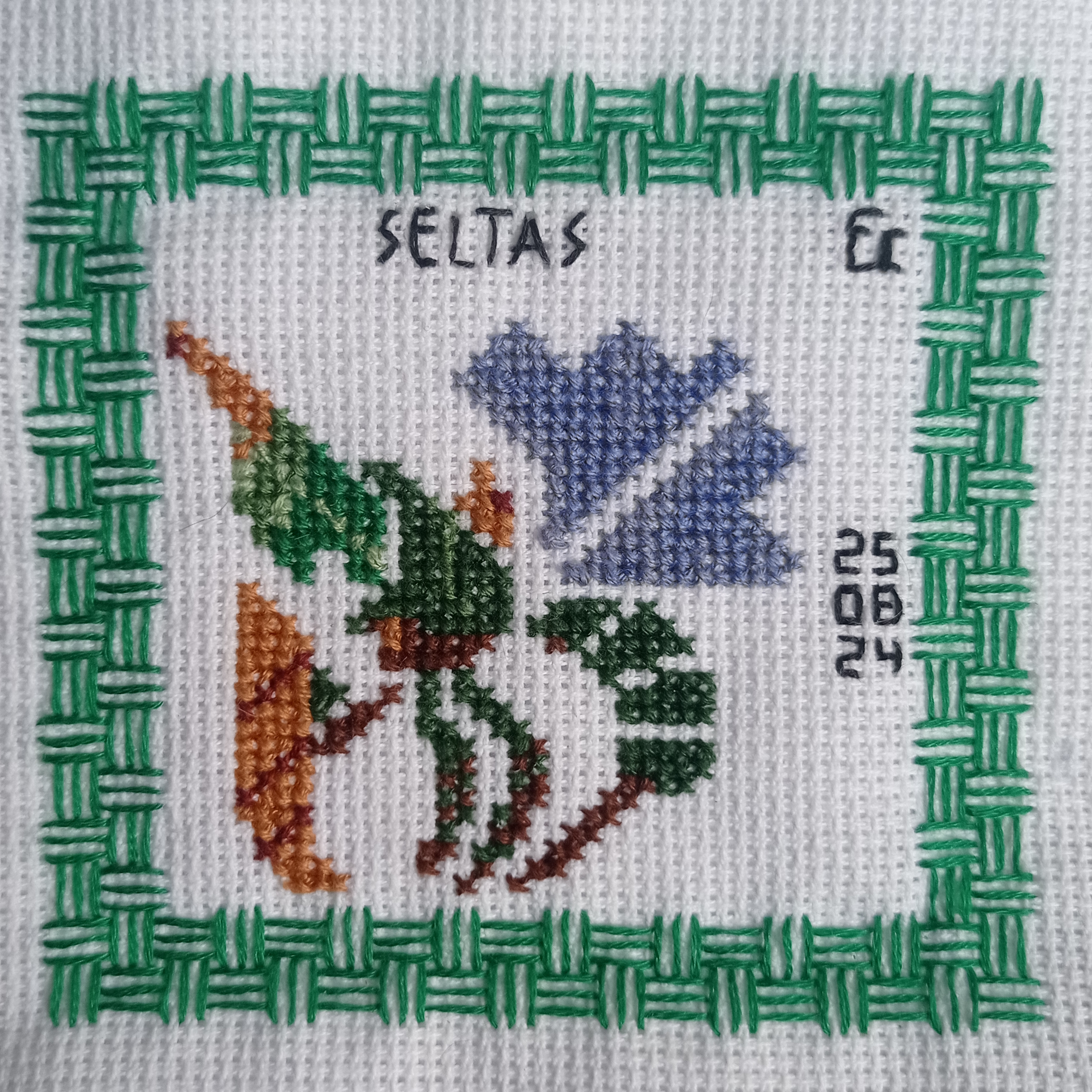 this is a closeup image of a cross stitch piece depicting a stylized flower in shades of green, blue and brown. the flower is framed by a border of green, and there is a date written on the right side of the frame, 25 08 24. the top of the frame also has the word "seltas" written.